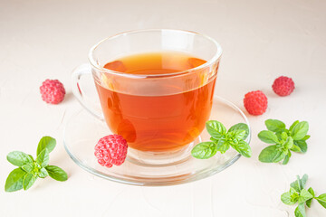 Herbal tea in a transparent cup with a saucer on a light background. Around - fresh raspberries and mint leaves. Vitamin drinks concept.