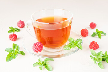 Herbal tea in a transparent cup with a saucer on a light background. Around - fresh raspberries and mint leaves. Vitamin drinks concept.