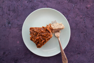 piece of pie covered with caramel and sunflower seeds in light blue plate with fork on a violet grunge background
