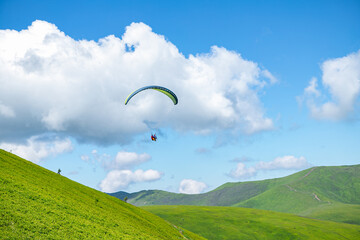 High view mountain panorama in the Carpathians. Two people on a paraglider. Paraglider fly over a mountain valley on a sunny summer day. Beautiful landscape with greenery and clouds.