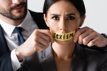 bearded businessman touching scotch tape with sexism lettering on mouth of businesswoman looking at...