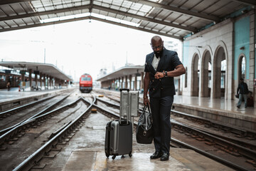 Stylish dark-skinned bald bearded businessman in glasses and an elegant suit is late for a business trip and looking at the wristwatch while standing on the railway platform inside the indoor station