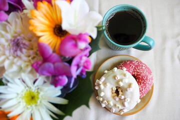two doughnuts with pink icing and white on a brown round plate with coffee in a turquoise mug and a bouquet of bright summer flowers on a beige background top view