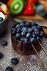 In focus blueberries with out of focus chopping board with knife and fresh fruit with fruit salad in bowl to the side.