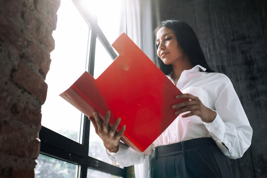 Mexican businesswoman holding a red open folder for documents next to a panoramic window in the loft