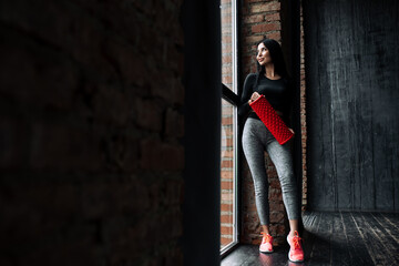 Full-length portrait of a fitness trainer in tight leggings leaning on a wall holding a massage roller and looking at the panoramic window in the loft