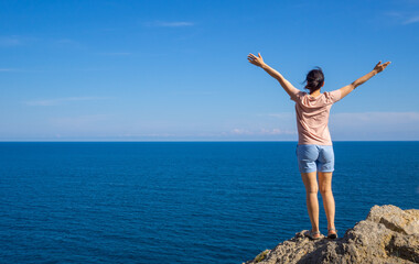 woman brunette in t-shirt stands back on rock, hands out to side, and looks out to sea. Solo summer outdoor activities. Hiking and trekking concept. Copy space