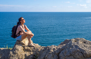 Fototapeta na wymiar brunette woman in t-shirt with backpack sits back on rocky seaside and looks out at the sea. Solo summer outdoor activities in fresh air. Concept of Hiking and trekking. Copy space