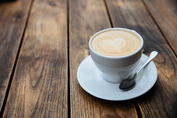 White ceramic cup of freshly brewed coffee with latte art in shape of heart. Hot beverage served on saucer with shiny spoon. Dark wooden background.