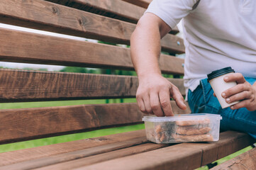 The guy sits on a bench and dines with a toast with a coffee in a glass. Fast food in the fresh air.