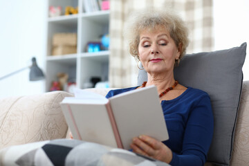 Portrait of well-educated senior female reading interesting story book. Attractive woman sitting on comfort couch at home and relaxing. Cozy interior. Hobby and spare time concept