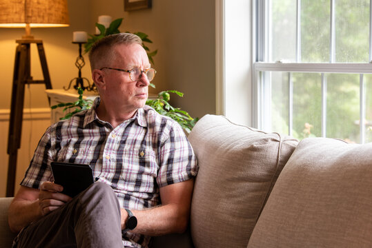 Mature man, sitting alone inside with a tablet, looks out living room window on a rainy day.