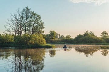 Summer landscape in Belarus. Colorful sky and reflections in the river at dawn. Man, a fisherman on a rubber boat in the middle of the river.