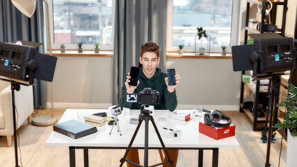 Time to compare. Male technology blogger recording video blog or vlog review of new smartphone, showing it and another one on camera at home studio. Blogging, Work from Home concept