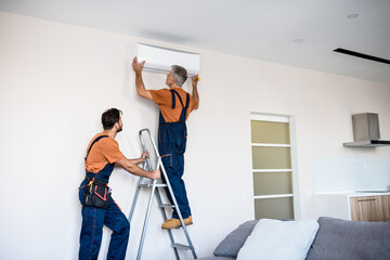 Exceptional service. Two workers in uniform, air conditioning masters using ladder while installing a new air conditioner in the apartment. Construction, maintenance and repair concept