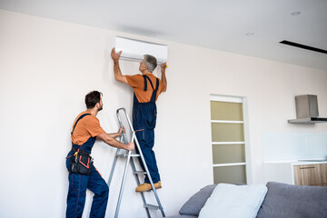 Exceptional service. Two workers in uniform, air conditioning masters using ladder while installing...