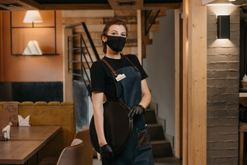 Fototapeta na wymiar A waitress who wears a medical face mask and black disposable medical gloves is posing with a plastic tray in a restaurant. A cute barista is waiting for clients in a cafe.