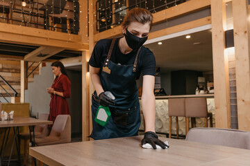 A cute waitress who wears a black medical face mask and disposable medical gloves is holding bottle with sanitizer and cleaning tables with rag in a restaurant. Staff is waiting for clients in a cafe.