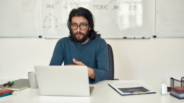 Learn Computer Programming online. Young focused bearded male teacher teaching computer science online through video call while sitting against whiteboard