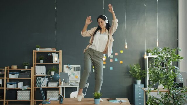 Happy businesswoman is dancing on office desk fooling around enjoying music in headphones in workplace. People, lifestyle and workspace concept.