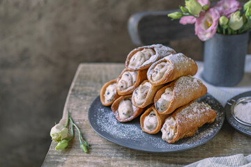 Sicilian cannoli deep fried pastry tubes on wooden rustic table with a sweet ricotta cream and dried candied fruits. Homemade Italian dessert on blue plate with flowers. Copy space, selective focus.