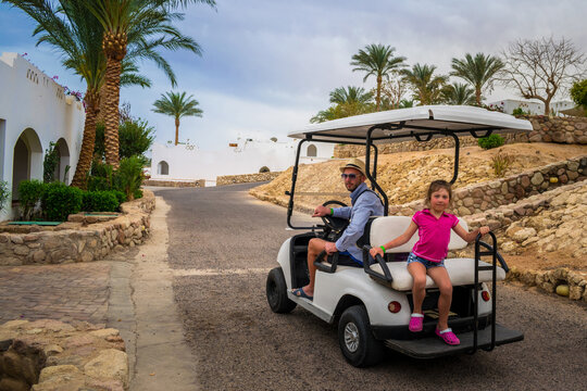 A man rides a little girl on a golf cart. The driver carries the child through a beautiful garden at a walking electric car.