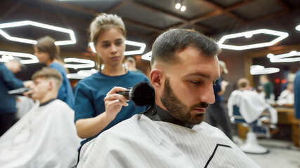 Obraz premium Young professional barber girl cleaning neck of a handsome bearded man after hair cutting. People visiting barbershop or beauty salon