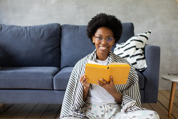 Happy African American young woman sit relax on cozy couch and reads a book. Happy to move to new...