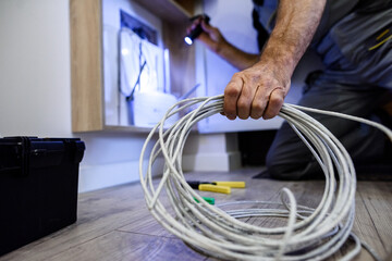 Close up shot of hand of aged electrician, repairman in uniform working, fixing, installing an ethernet cable in fuse box, holding flashlight and cable - obrazy, fototapety, plakaty