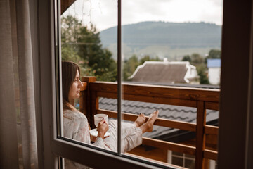 Young female standing after taking a shower in the morning on balcony of the hotel. holding a cup of coffee or tea in her hands. Looking outside nature forest and Mountain