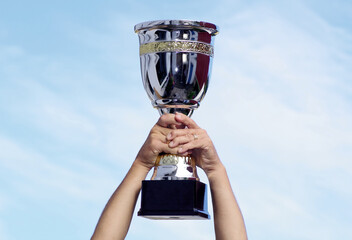 Winner takes it all. Trophy cup against the sky, held by two hands.