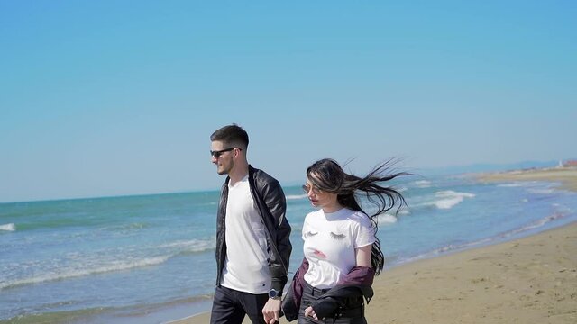 Happy young couple guy and girl in black clothes with black hair in sunglasses walking embracing having fun by the sea. Stylish fashion woman and man holding hands together. Ocean beach sand nature