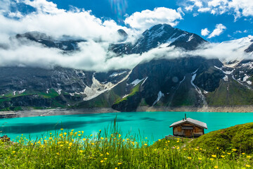 Mooserboden alpine lake in the Austria alps with wooden house and low clouds and blooming flowers in front