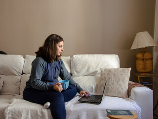 young woman sitting on a white sofa working on a laptop holding a coffee mug using comfort clothes 