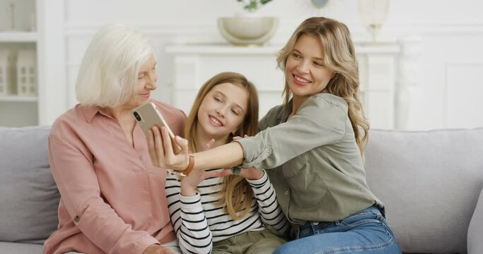 Caucasian pretty teen girl sitting on couch with mother and grandmother, posing to smartphone camera and taking selfie photos. Senior woman with daughter and granddaughter at home. Phone photo making.