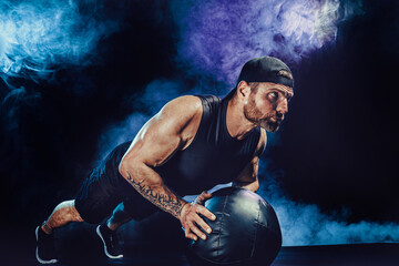 Aggressive bearded muscular sportsman is working out, push up with a medicine ball isolated on dark studio background with smoke.