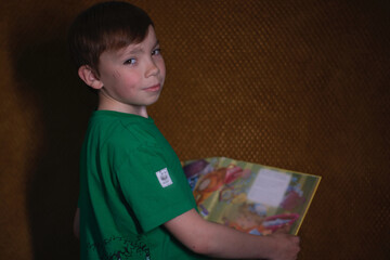 portrait of a pensive nine year old blond boy in a green t-shirt on brown background reading a yellow book with bright pictures. home schooling 