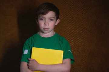 portrait of a pensive nine year old blond boy in a green t-shirt on brown background holding a yellow book. Home schooling. 