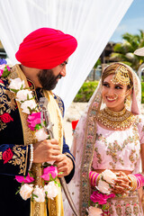 Sikh bride and groom wearing bright traditional clothing on clear sandy beach beneath sunny blue skies