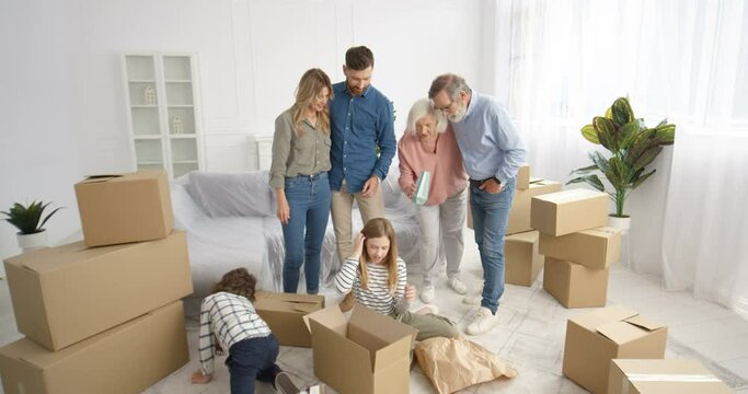 Caucasian parents and grandparents in living room with small cute kids unpacking carton boxes. Family with children and elderly moving in new house after repairment. Replacing in new apartment.