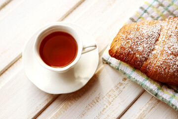 A white Cup and saucer with black tea and a fresh soft delicious croissant sprinkled with powdered sugar on a textile napkin on a vintage wooden surface. French breakfast. Selective focus
