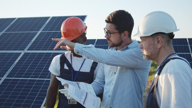Multi-ethnic electrician technicians talking with engineer about installation of ecological solar panels on field. Renewable energy plant. Ecology. Teamwork.