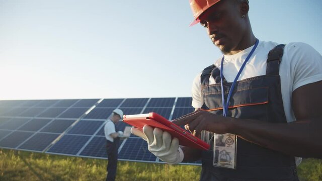 Black Energy Specialist Using Digital Tablet Reading Information To Check The Efficiency Of Solar Panel Construction. Green Energy Jobs. Technology. Innovation.