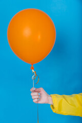 Female hand in yellow jacket holding orange balloon, photo on blue background. Happy air flying ball
