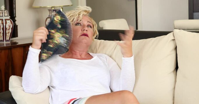 Portrait of a middle aged woman suffering a heat wave using a fan sitting on a couch in the living room at home