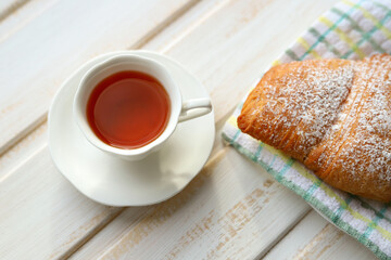 A white Cup and saucer with black tea and a fresh soft delicious croissant sprinkled with powdered sugar on a textile napkin on a vintage wooden surface. French breakfast. Selective focus breakfast. 