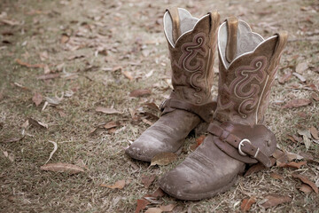 pair of dirty old cowboy boots with spur straps on a background of dry grass and leaves.