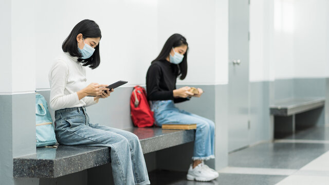 Two female college students wearing mask, reading book and learning while keeping social distance in university campus during COVID-19 pandemic. Stock photo.