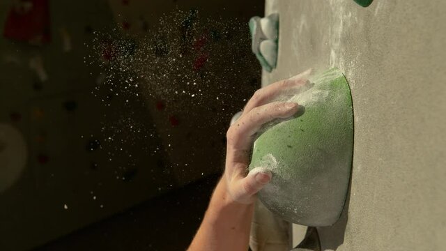 SLOW MOTION, MACRO, DOF: Detailed shot of a green sloper hold as unrecognizable climber grips it with her magnesium-covered hand. Tiny chalk particles fly in the air as rock climber trains indoors.