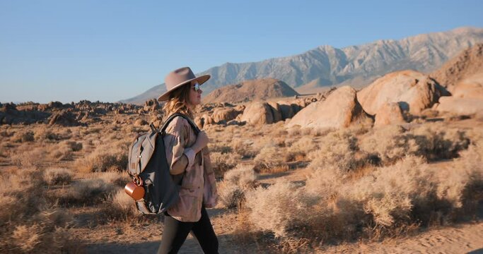 Camera follows millennial young woman walking by the wild desert landscape at sunrise. Smiling model in fashionable ranchero hat, camouflage jacket and backpack is hiking at beautiful nature park, USA